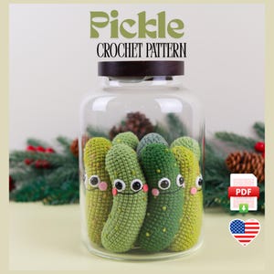 Op de afbeelding: Een haakpatroon voor het maken van amigurumi-augurken. Het patroon is beschikbaar als PDF-download. De afbeelding toont een glazen pot gevuld met verschillende groene gehaakte augurken met glimlachende gezichten.