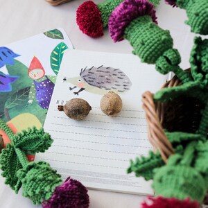 Pode incluir: Um close-up de uma flor de cardo de croch&ecirc; feita &agrave; m&atilde;o com um caule verde e um pompom roxo. A flor est&aacute; sobre uma superf&iacute;cie branca ao lado de um caderno com uma ilustra&ccedil;&atilde;o de ouri&ccedil;o e a palavra "NO" escrita nele.