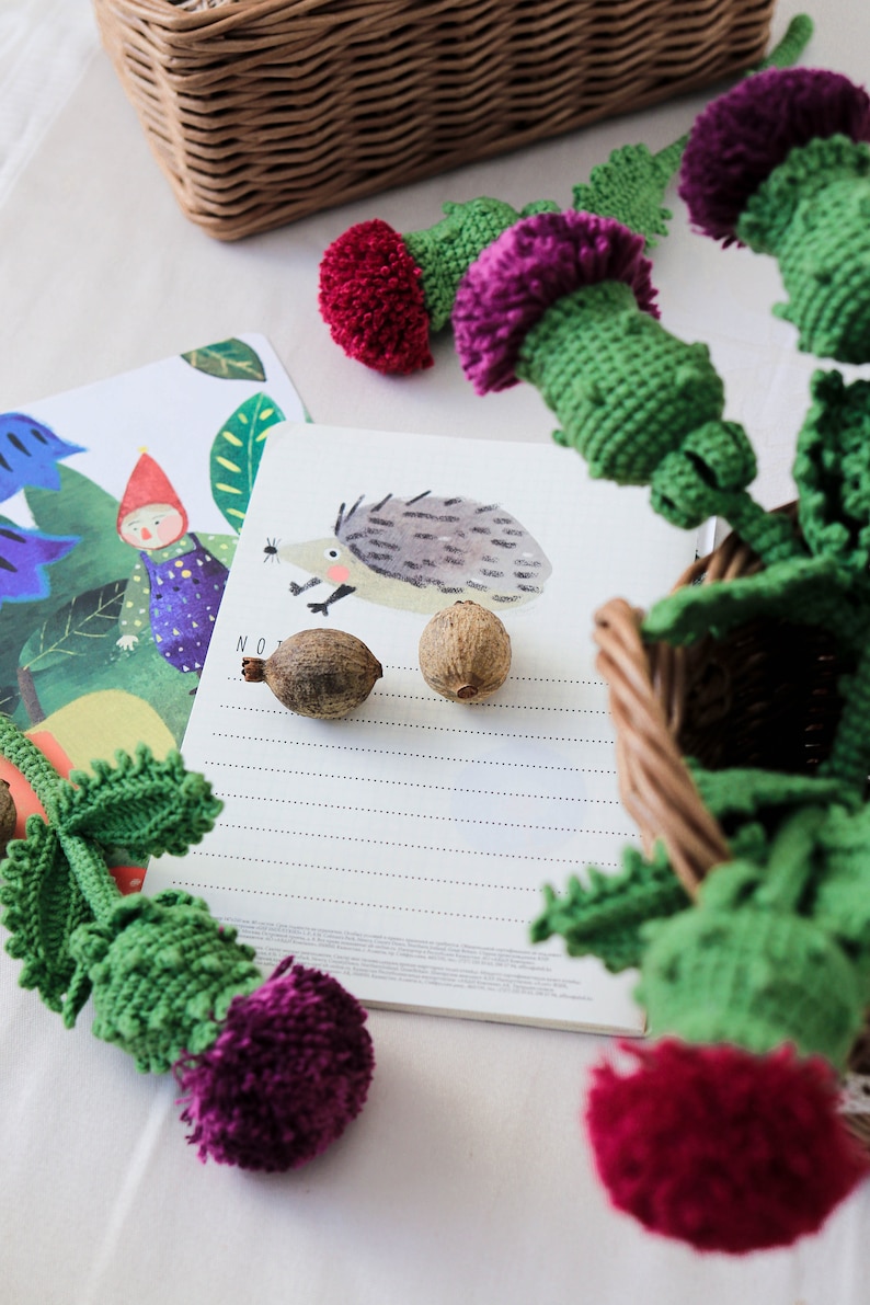 Pode incluir: Um close-up de uma flor de cardo de croch&ecirc; feita &agrave; m&atilde;o com um caule verde e um pompom roxo. A flor est&aacute; sobre uma superf&iacute;cie branca ao lado de um caderno com uma ilustra&ccedil;&atilde;o de ouri&ccedil;o e a palavra "NO" escrita nele.