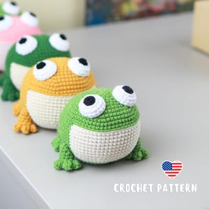 May include: Crocheted frog toys in green, yellow, and pink, with white bellies and large, expressive eyes. The toys are arranged in a row, showcasing the handmade details. The text "CROCHET PATTERN" is visible.