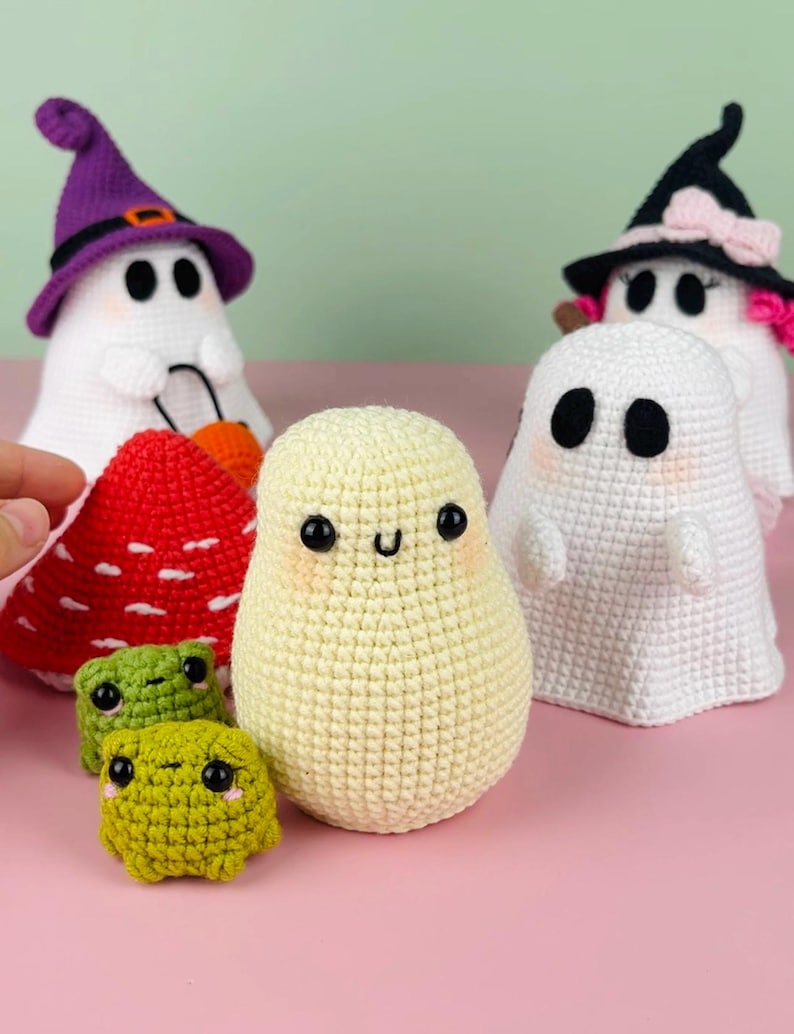 Puede incluir: Un grupo de juguetes de crochet con tem&aacute;tica de Halloween, que incluyen fantasmas, un hongo y dos ranas verdes. Los fantasmas llevan sombreros de bruja y tienen caras lindas. El hongo es rojo con manchas blancas. Las ranas son verdes con ojos negros y caras sonrientes.