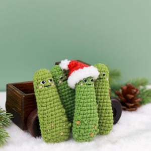 May include: A group of four green crocheted pickle ornaments with smiling faces. One pickle wears a red and white Santa hat. The pickles are arranged in front of a small wooden wagon on a white surface, with a green background.