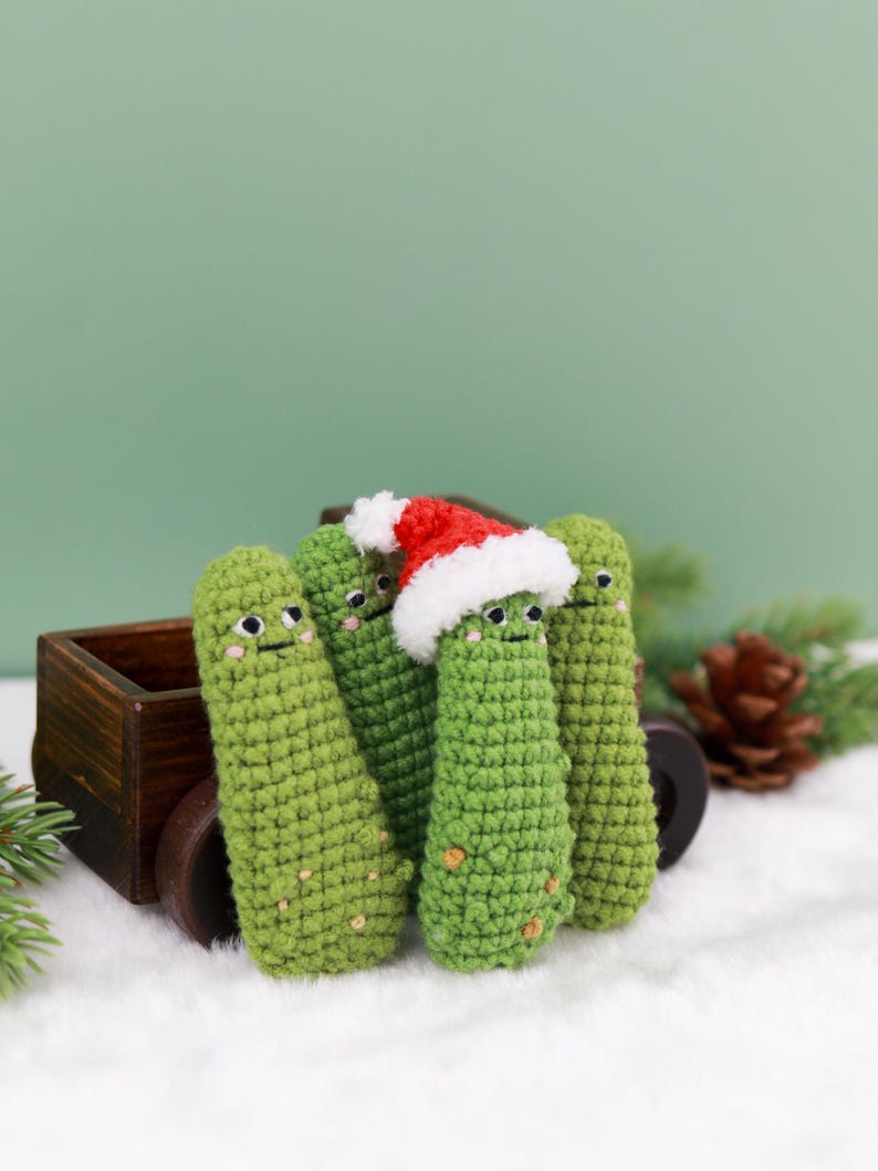 May include: A group of four green crocheted pickle ornaments with smiling faces. One pickle wears a red and white Santa hat. The pickles are arranged in front of a small wooden wagon on a white surface, with a green background.