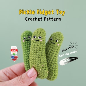May include: Three green crocheted pickle fidget toys with smiling faces. The text "Pickle Fidget Toy Crochet Pattern" is at the top. A hair clip is included. The image also includes a PDF icon and an American flag.