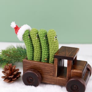 May include: A wooden toy truck carries four green crocheted figures, one wearing a Santa hat. A pine cone and evergreen sprig are in the foreground. The figures have small black eyes. The truck is brown.
