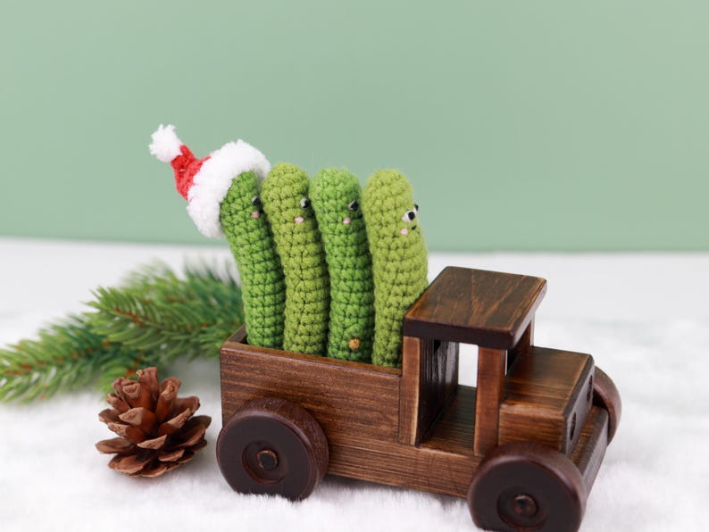 May include: A wooden toy truck carries four green crocheted figures, one wearing a Santa hat. A pine cone and evergreen sprig are in the foreground. The figures have small black eyes. The truck is brown.
