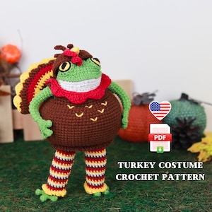 May include: Crochet pattern for a turkey costume for a frog. The pattern is available as a PDF download. The frog is wearing a brown turkey costume with a red and yellow feathered tail. The frog has green skin and yellow eyes.