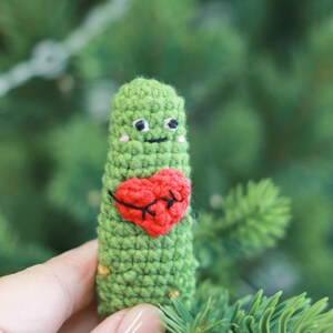 May include: A handmade green crochet figure with a red heart applique. The figure has a smiling face with black eyes and a mouth. The figure is held in front of a blurred green background.