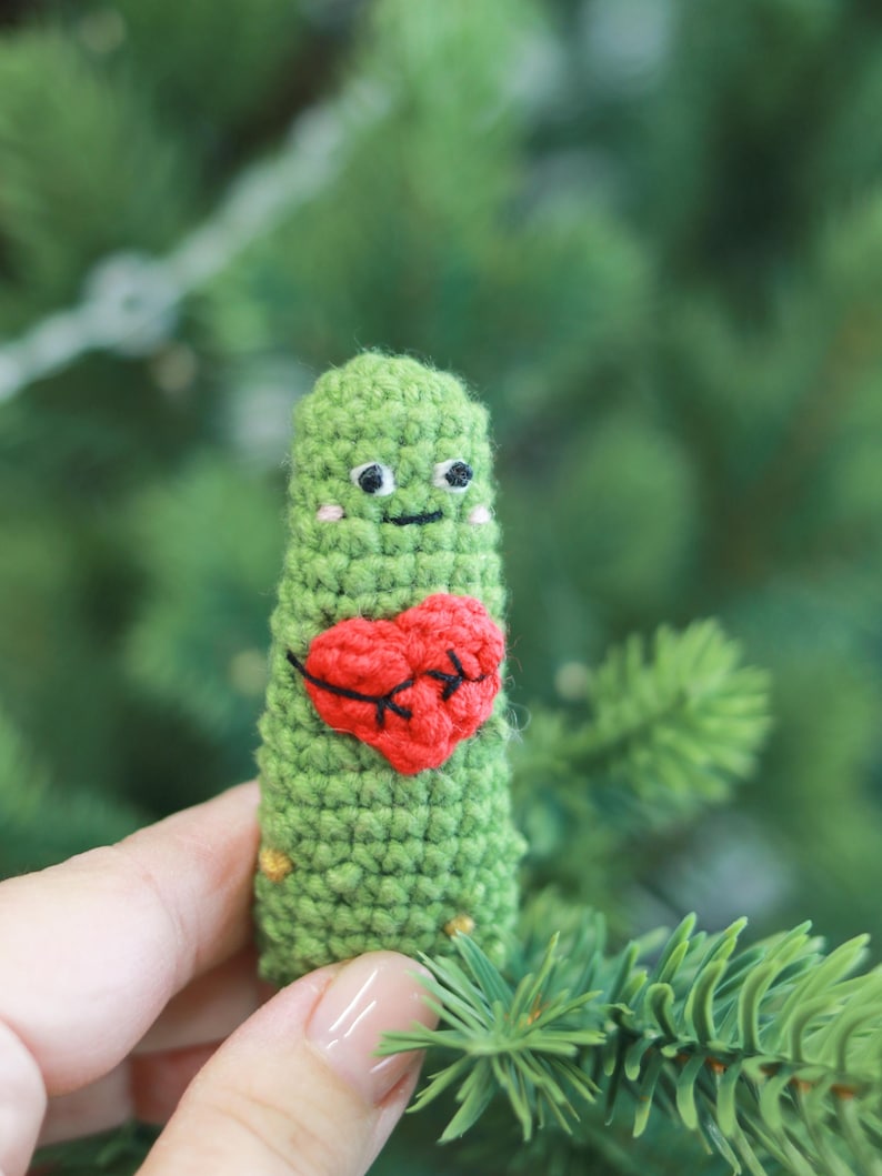 May include: A handmade green crochet figure with a red heart applique. The figure has a smiling face with black eyes and a mouth. The figure is held in front of a blurred green background.