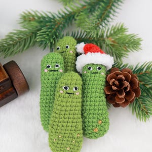 May include: A group of four green crocheted pickle-shaped ornaments with smiling faces. One pickle wears a red and white Santa hat. The ornaments are arranged with pine needles and a pine cone on a white surface.