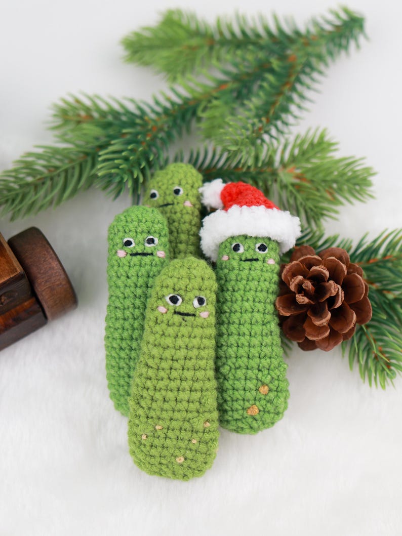 May include: A group of four green crocheted pickle-shaped ornaments with smiling faces. One pickle wears a red and white Santa hat. The ornaments are arranged with pine needles and a pine cone on a white surface.