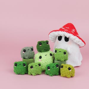 An adorable ghost wearing a red-spotted toadstool hat, cradling a tiny green frog in its hands. The ghost features black felt eyes, rosy embroidered cheeks, and a playful surprise—under its white cloak hides a hidden potato man! A whimsical handmade crochet creation full of sweet details. Perfect for Halloween decor.