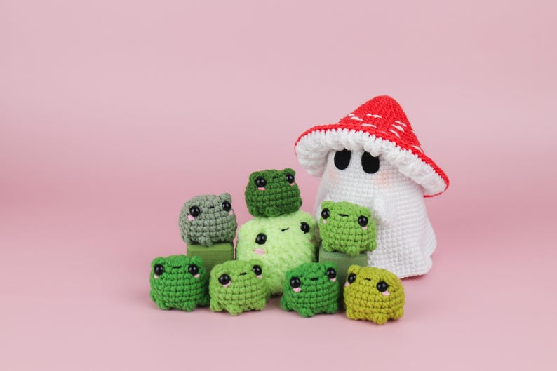 An adorable ghost wearing a red-spotted toadstool hat, cradling a tiny green frog in its hands. The ghost features black felt eyes, rosy embroidered cheeks, and a playful surprise—under its white cloak hides a hidden potato man! A whimsical handmade crochet creation full of sweet details. Perfect for Halloween decor.