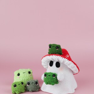 An adorable ghost wearing a red-spotted toadstool hat, cradling a tiny green frog in its hands. The ghost features black felt eyes, rosy embroidered cheeks, and a playful surprise—under its white cloak hides a hidden potato man! A whimsical handmade crochet creation full of sweet details. Perfect for Halloween decor.