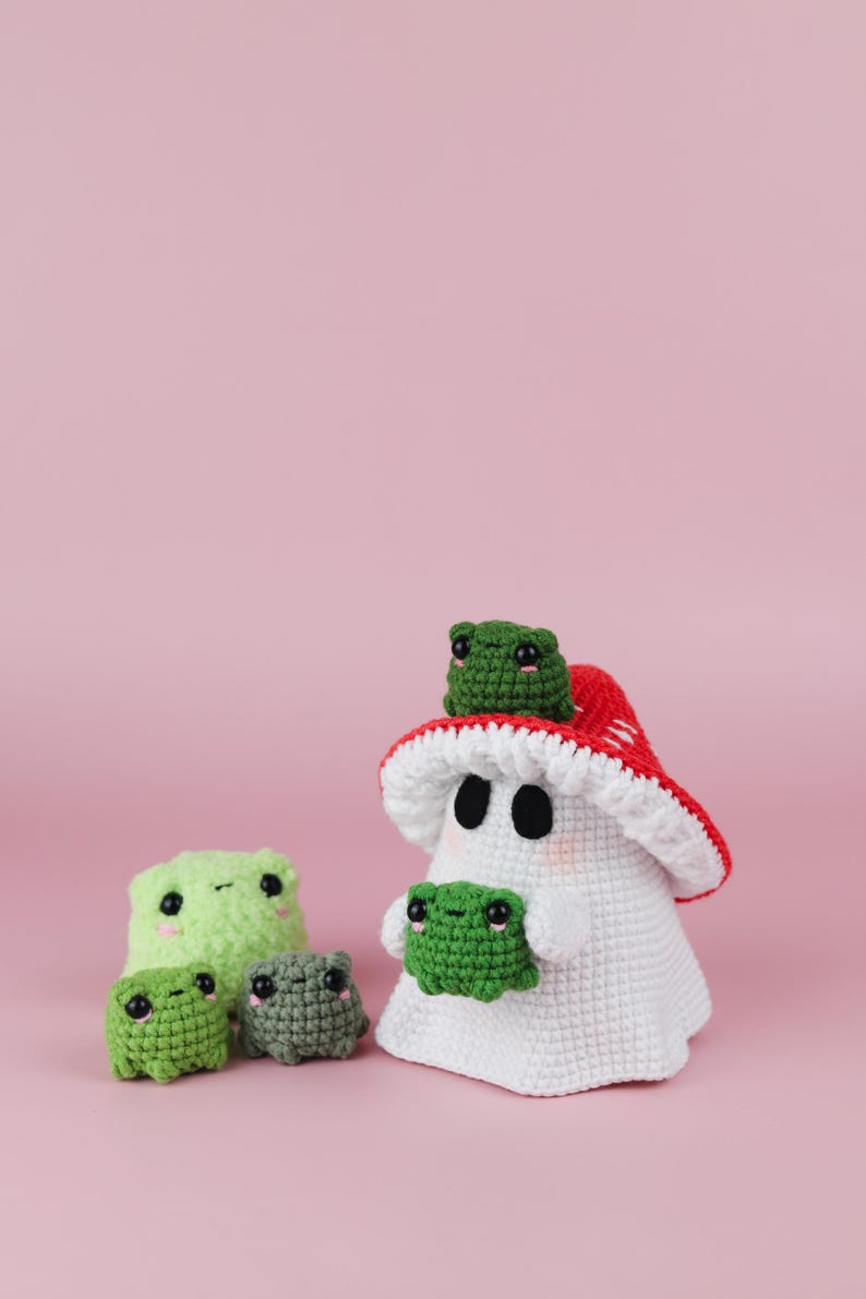 An adorable ghost wearing a red-spotted toadstool hat, cradling a tiny green frog in its hands. The ghost features black felt eyes, rosy embroidered cheeks, and a playful surprise—under its white cloak hides a hidden potato man! A whimsical handmade crochet creation full of sweet details. Perfect for Halloween decor.