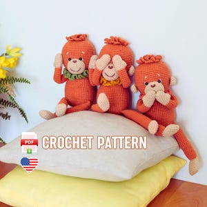 Może przedstawiać: Trzy pomarańczowe lalki małpki na szydełku, każda w innej pozie: zakrywająca uszy, oczy i usta. Siedzą na białej poduszce nad żółtą poduszką. Na zdjęciu widnieje napis "CROCHET PATTERN".