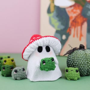 An adorable ghost wearing a red-spotted toadstool hat, cradling a tiny green frog in its hands. The ghost features black felt eyes, rosy embroidered cheeks, and a playful surprise—under its white cloak hides a hidden potato man! A whimsical handmade crochet creation full of sweet details. Perfect for Halloween decor.