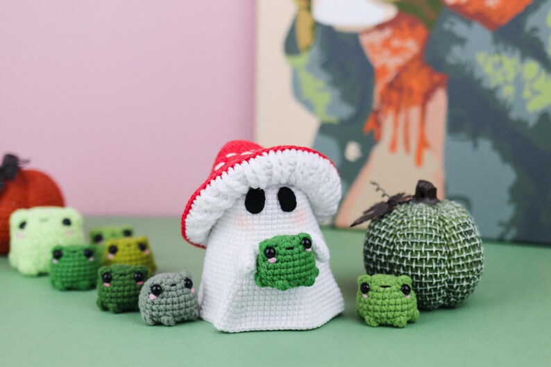 An adorable ghost wearing a red-spotted toadstool hat, cradling a tiny green frog in its hands. The ghost features black felt eyes, rosy embroidered cheeks, and a playful surprise—under its white cloak hides a hidden potato man! A whimsical handmade crochet creation full of sweet details. Perfect for Halloween decor.