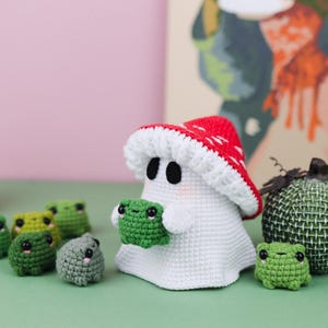 An adorable ghost wearing a red-spotted toadstool hat, cradling a tiny green frog in its hands. The ghost features black felt eyes, rosy embroidered cheeks, and a playful surprise—under its white cloak hides a hidden potato man! A whimsical handmade crochet creation full of sweet details. Perfect for Halloween decor.