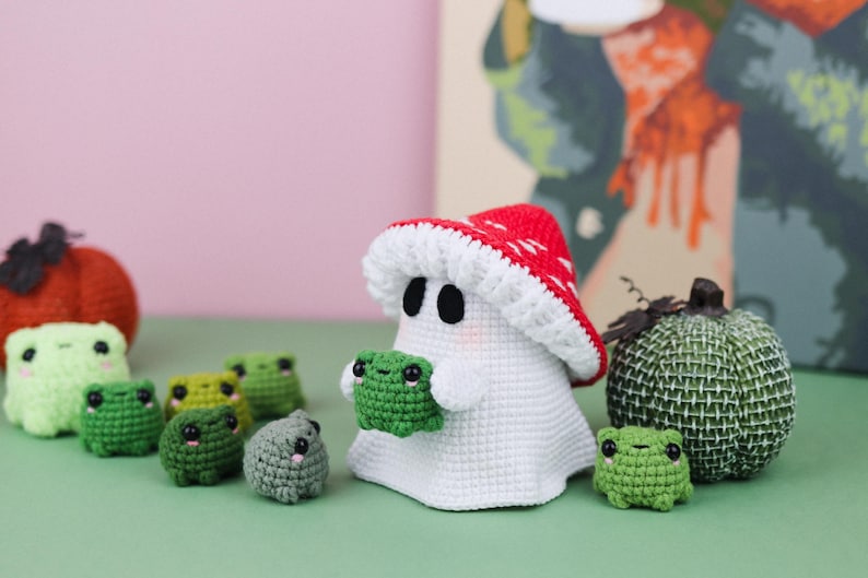 An adorable ghost wearing a red-spotted toadstool hat, cradling a tiny green frog in its hands. The ghost features black felt eyes, rosy embroidered cheeks, and a playful surprise—under its white cloak hides a hidden potato man! A whimsical handmade crochet creation full of sweet details. Perfect for Halloween decor.