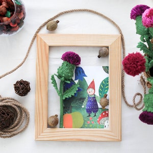 Pode incluir: Uma moldura de madeira com um passe-partout branco e uma ilustra&ccedil;&atilde;o colorida de um gnomo e um cardo roxo. A moldura est&aacute; sobre uma superf&iacute;cie branca com uma corda marrom e uma pequena bola marrom.