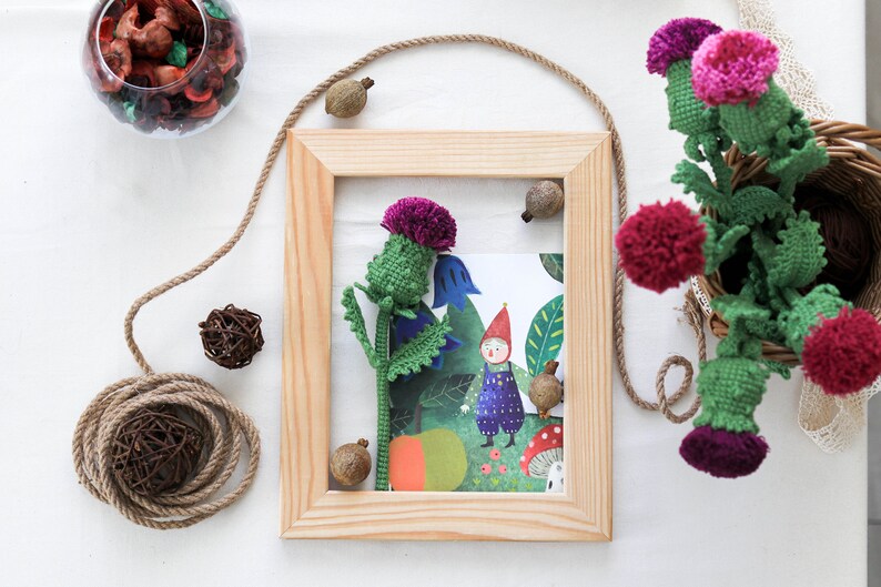 Pode incluir: Uma moldura de madeira com um passe-partout branco e uma ilustra&ccedil;&atilde;o colorida de um gnomo e um cardo roxo. A moldura est&aacute; sobre uma superf&iacute;cie branca com uma corda marrom e uma pequena bola marrom.