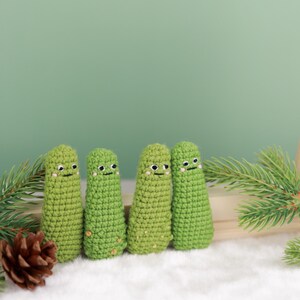 May include: Four green crocheted figures with smiling faces and small brown accents. The figures are arranged in a row on a white surface, with pine needles and a pine cone in the background. The figures have a whimsical, handmade appearance.