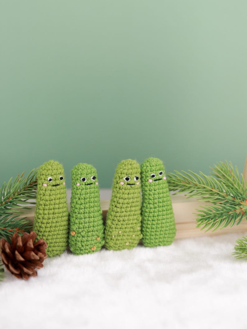 May include: Four green crocheted figures with smiling faces and small brown accents. The figures are arranged in a row on a white surface, with pine needles and a pine cone in the background. The figures have a whimsical, handmade appearance.