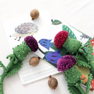 Pode incluir: Tr&ecirc;s flores de cardo de croch&ecirc; com caules verdes e pompons roxos e vermelhos. As flores est&atilde;o sobre uma superf&iacute;cie branca com um caderno e algumas bolotas marrons.