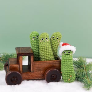 May include: A wooden toy truck carries four green crocheted pickle figures with smiling faces. One pickle wears a Santa hat. The truck sits on a bed of white faux snow, with a green background and pine branches.