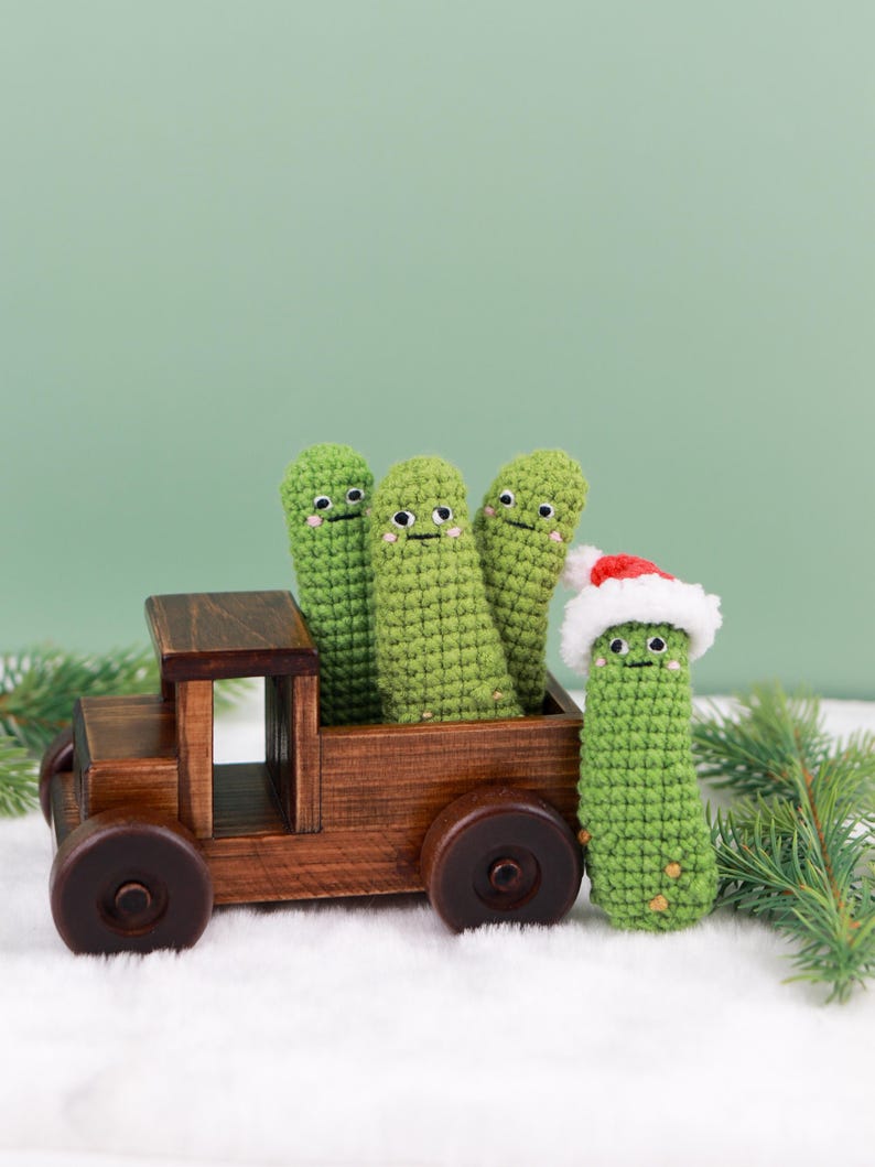 May include: A wooden toy truck carries four green crocheted pickle figures with smiling faces. One pickle wears a Santa hat. The truck sits on a bed of white faux snow, with a green background and pine branches.