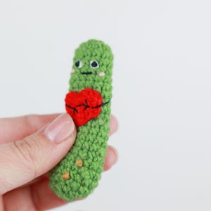 May include: A handmade green crochet pickle with a red heart. The pickle has embroidered black eyes, a smile, and blush. The heart is stitched onto the pickle with black thread. The pickle is held in a hand against a white background.
