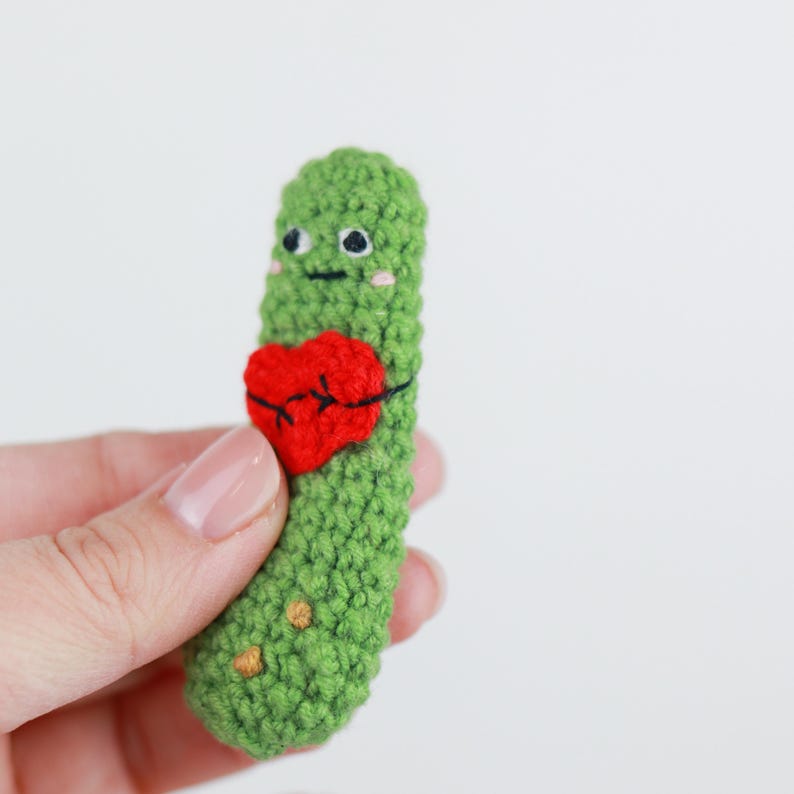 May include: A handmade green crochet pickle with a red heart. The pickle has embroidered black eyes, a smile, and blush. The heart is stitched onto the pickle with black thread. The pickle is held in a hand against a white background.
