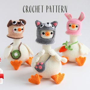 Könnte beinhalten: Drei gehäkelte Amigurumi-Spielzeug-Gänse, die verschiedene Tierhüte tragen. Die Gans links trägt einen braunen und weißen Mops-Hut, die Gans in der Mitte trägt einen grauen und weißen Katzenhut und die Gans rechts trägt einen rosa Hasenhut. Die Gänse sind alle weiß mit orangenen Füßen und Schnäbeln.