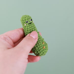 May include: A handmade, green crocheted pickle with a smiling face and small orange accents. The pickle is held in a hand against a light green background. The crocheted pickle is approximately 3 inches long.