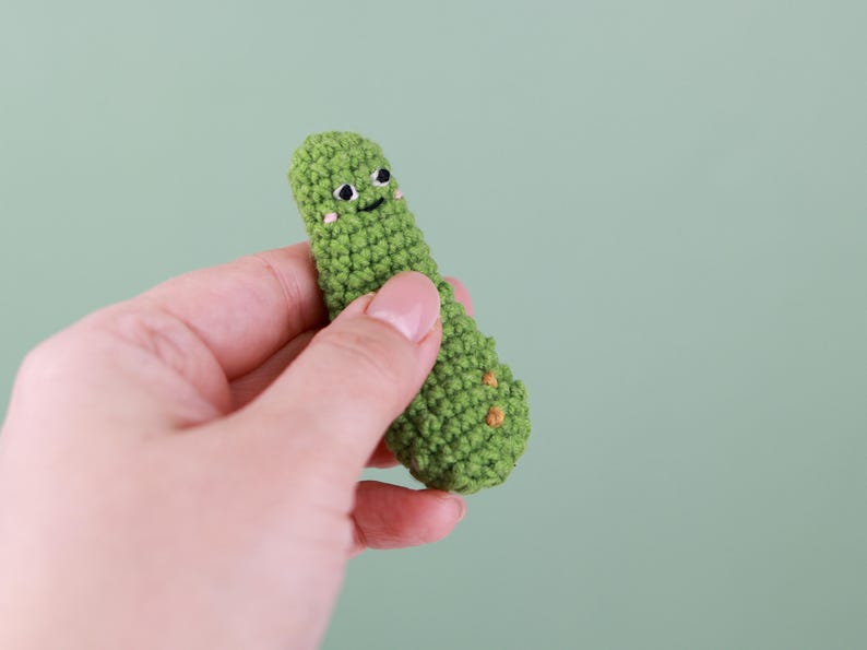 May include: A handmade, green crocheted pickle with a smiling face and small orange accents. The pickle is held in a hand against a light green background. The crocheted pickle is approximately 3 inches long.