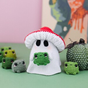 An adorable ghost wearing a red-spotted toadstool hat, cradling a tiny green frog in its hands. The ghost features black felt eyes, rosy embroidered cheeks, and a playful surprise—under its white cloak hides a hidden potato man! A whimsical handmade crochet creation full of sweet details. Perfect for Halloween decor.
