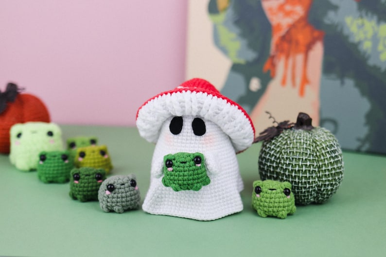 An adorable ghost wearing a red-spotted toadstool hat, cradling a tiny green frog in its hands. The ghost features black felt eyes, rosy embroidered cheeks, and a playful surprise—under its white cloak hides a hidden potato man! A whimsical handmade crochet creation full of sweet details. Perfect for Halloween decor.