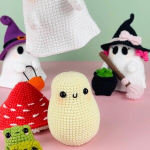 Puede incluir: Una colecci&oacute;n de juguetes de ganchillo con tem&aacute;tica de Halloween, que incluye un fantasma, una bruja, un hongo y dos ranas. Los juguetes est&aacute;n hechos con hilo blanco, verde, rojo y morado.
