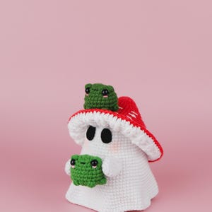 An adorable ghost wearing a red-spotted toadstool hat, cradling a tiny green frog in its hands. The ghost features black felt eyes, rosy embroidered cheeks, and a playful surprise—under its white cloak hides a hidden potato man! A whimsical handmade crochet creation full of sweet details. Perfect for Halloween decor.