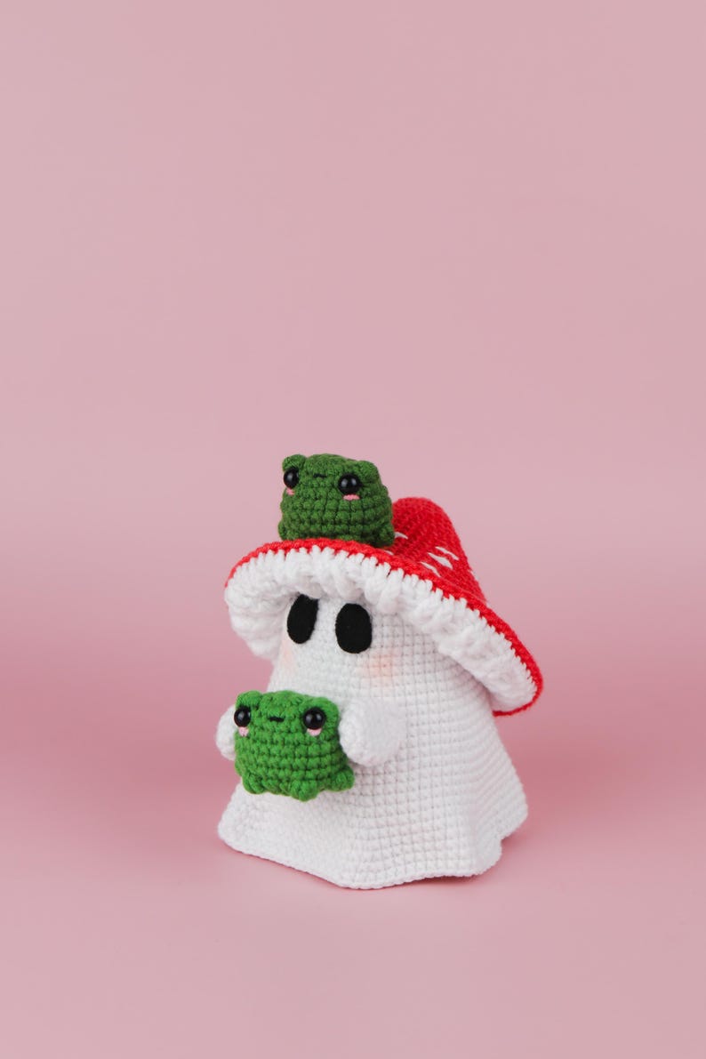 An adorable ghost wearing a red-spotted toadstool hat, cradling a tiny green frog in its hands. The ghost features black felt eyes, rosy embroidered cheeks, and a playful surprise—under its white cloak hides a hidden potato man! A whimsical handmade crochet creation full of sweet details. Perfect for Halloween decor.