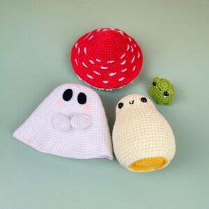 Puede incluir: Un conjunto de cuatro juguetes amigurumi de crochet: un fantasma blanco, un huevo amarillo, una seta roja y blanca y una rana verde.