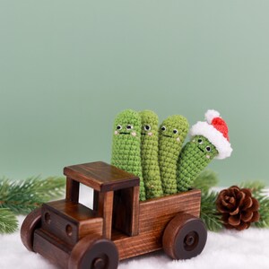 May include: A wooden toy truck carries four green crocheted figures with smiling faces. One figure wears a Santa hat. The truck sits on a bed of white fluff with a pine cone and evergreen sprig in the background. The truck is made of dark brown wood.