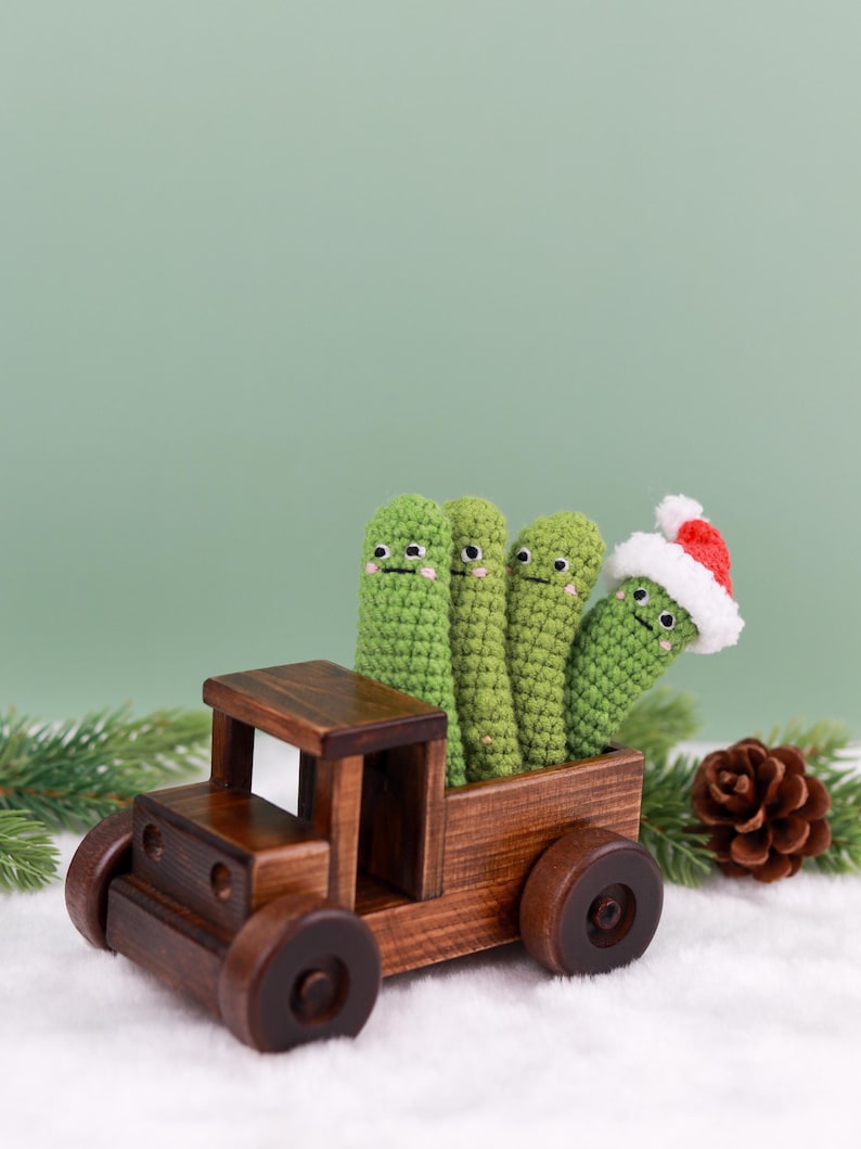 May include: A wooden toy truck carries four green crocheted figures with smiling faces. One figure wears a Santa hat. The truck sits on a bed of white fluff with a pine cone and evergreen sprig in the background. The truck is made of dark brown wood.