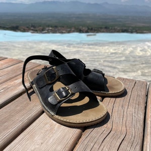 May include: A pair of black leather sandals with two adjustable buckles. The sandals have a brown sole and are sitting on a wooden surface.