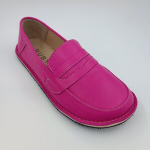 Peut inclure: Un mocassin en cuir rose vif avec un d&eacute;tail cousu sur le devant. Le mocassin a une semelle en caoutchouc et le mot "RUBY" est visible &agrave; l'int&eacute;rieur de la chaussure.