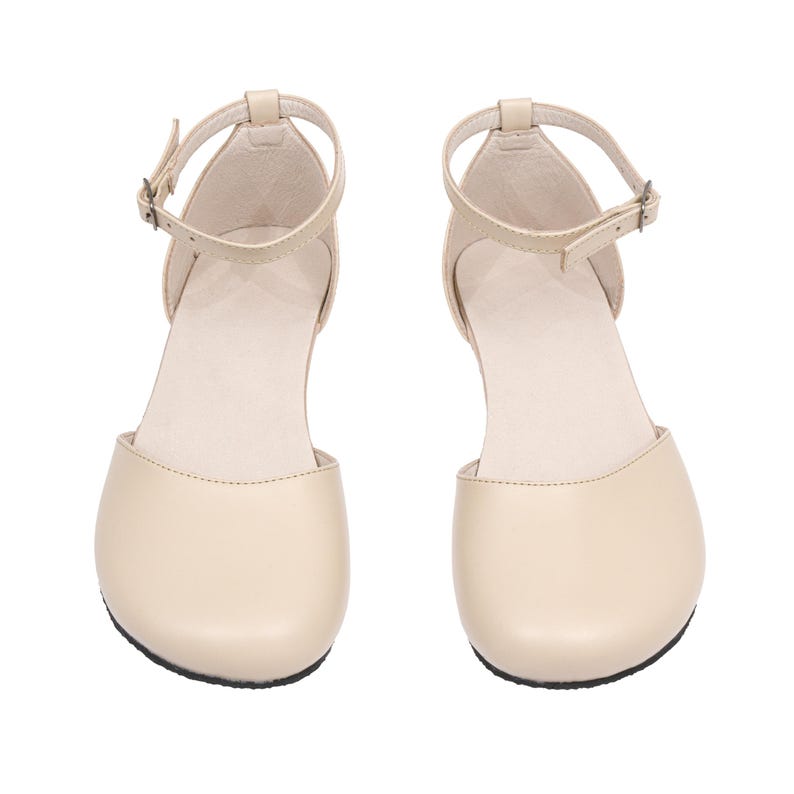 Puede incluir: Un par de sandalias de cuero beige con correas en el tobillo y peque&ntilde;as hebillas. Las sandalias tienen una punta redondeada y un tac&oacute;n bajo. Las suelas son negras.