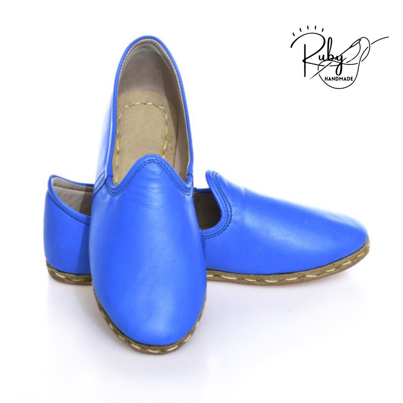 K&ouml;nnte beinhalten: Ein Paar blaue Lederschlappen mit braunem, gen&auml;hten Rand. Die Schuhe sind handgefertigt von Ruby.