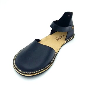 Puede incluir: Sandalias de cuero negro con una correa alrededor del tobillo. Las sandalias tienen un detalle de costura dorada alrededor de la suela y el nombre de la marca "RUBY barefoot" est&aacute; impreso en la plantilla.