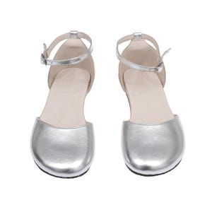 Puede incluir: Un par de bailarinas plateadas de punta cerrada con correas en el tobillo. Los zapatos tienen un brillo met&aacute;lico y un peque&ntilde;o cierre de hebilla. Las suelas son negras y el interior es de color beige claro. Los zapatos est&aacute;n dise&ntilde;ados para una ocasi&oacute;n formal o elegante.
